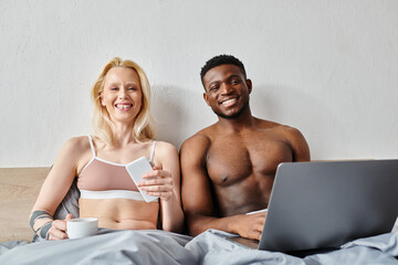 A multicultural couple sitting on a bed, engrossed in a laptop.
