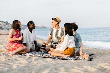 Young group of happy friends hanging out sitting on the beach enjoying summer vacation together