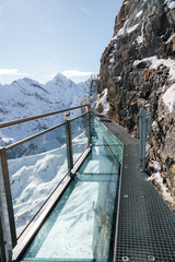 Glass floored cliff walkway with metal railings in Murren ski resort, Switzerland. Stunning alpine vista, snow covered peaks, and clear blue sky view.