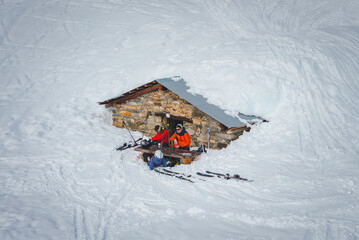 Alpine scene featuring small stone hut covered in snow, skiers taking a break. Winter sports concept with colorful gear. Snowy mountain slope setting.