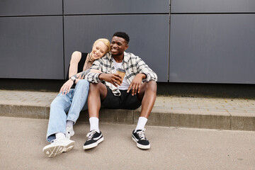 A man and woman sit peacefully on the ground by a grey building in an urban setting, enjoying each others company.