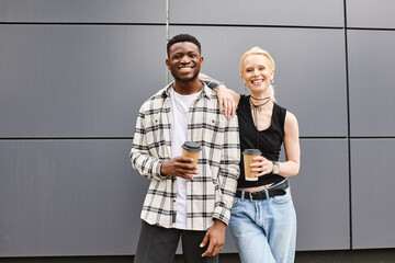 A multicultural couple stands happily side by side on an urban street near a grey building.