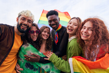 Happy portrait of a multiracial group of young people celebrating Gay Pride Festival Day with rainbow flags. LGBT community concept. Men and women hugging looking at camera outdoors. - obrazy, fototapety, plakaty