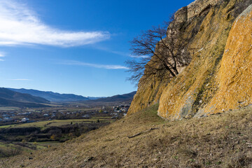View of the village and mountains. The rock is covered with yellow lichen. Tree without leaves.  Bright blue sky with clouds.  Kveshi fortress. Georgia