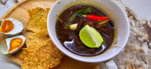 Rawon or Indonesian black beef soup, black color are from indonesian nut called kluwek. Served with lime, chili paste, salted egg, and tempe cracker. Perfect for recipe, article, or any cooking conten