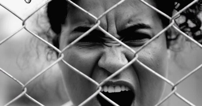 Desperate Young African American woman feeling outrage screaming with rage behind metal fence barrier frowning and open mouth screaming in anger