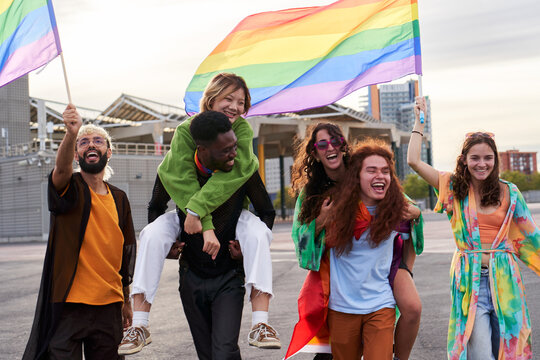 Excited people at a pride month festival, in piggybacks holding rainbow flags celebrating inclusion and LGBTQI rights happily. Multiethnic friends together in support of gay community.