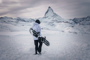 Snowboarder at Zermatt ski resort, facing Matterhorn mountain. Dressed in winter gear and holding a...