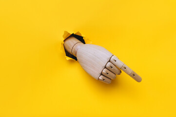 A wooden hand protrudes from a torn hole in yellow paper and points its index finger down to the right. Concept of direction, adjustment and assistance. Mannequin, robot.