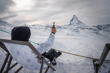 Winter scene at Zermatt ski resort person in snow gear holds up beer in deck chair overlooking Matterhorn mountain. Perfect blend of adventure and relaxation.