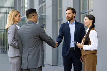 Two businessmen stand outside an office building together with female assistants, shake hands,...