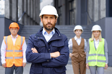 Portrait of serious young male engineer, owner in uniform and hard hat standing in front of camera...