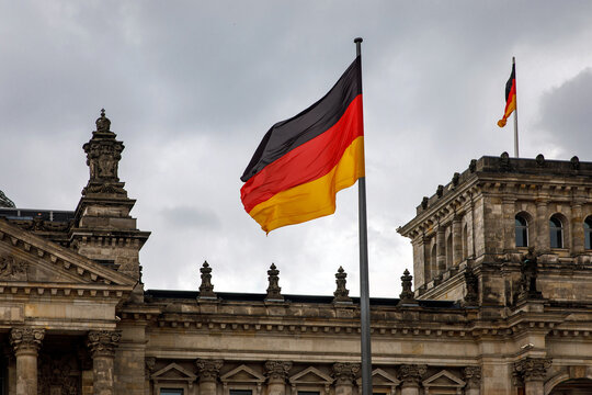 The flag of Germany flutters proudly near the German Parliament building against a cloudy sky.