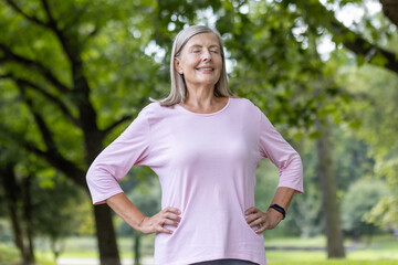 An older gray-haired woman stands in a park in a supportive outfit, holds her hands on the side of her body and closes her eyes, meditates, breathes relaxed.