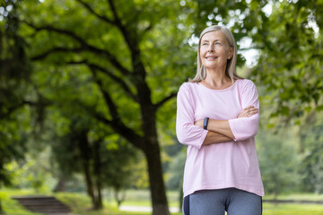 Photo of an older gray-haired smiling and relaxed woman standing in a park in casual clothes, clasping her hands on her chest and looking to the side.