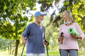 Senior smiling couple, gray-haired man and woman doing sports in the park. Standing holding trekking poles, mat and water, talking to each other.