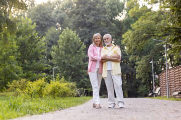 Fototapeta na wymiar Portrait of senior gray-haired couple, man and woman standing in casual clothes in park, hugging each other and looking at camera.