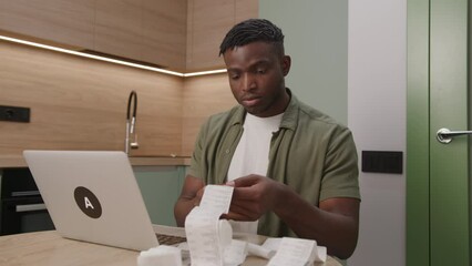 Young man examining receipts with a laptop on a kitchen counter