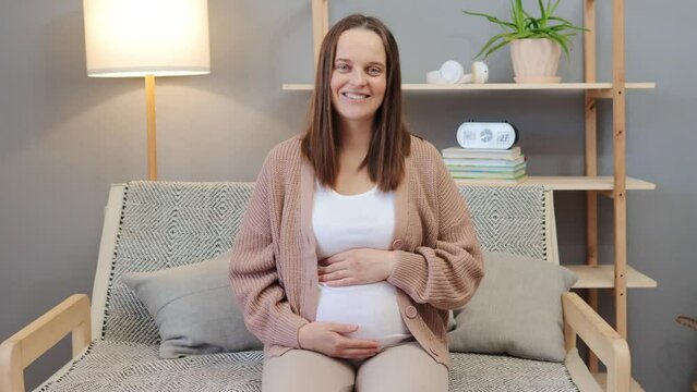 Joyful pregnant dark haired woman in beige jumper and white T-shirt sitting on sofa stroking her belly talking with psychotherapist having pleasant conversation about her feelings during pregnancy