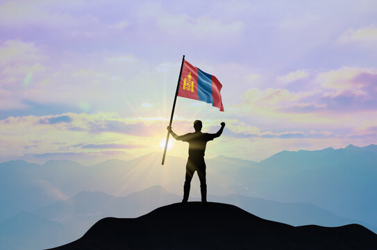 Mongolia flag being waved by a man celebrating success at the top of a mountain against sunset or sunrise. Mongolia flag for Independence Day.