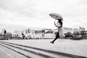 A girl in a light brown coat and a light scarf under an umbrella in a jump shot on a cloudy April evening against the background of Cheboksary