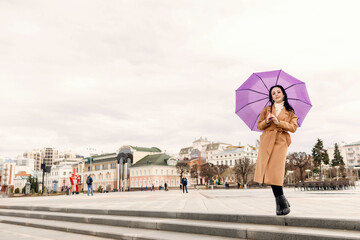 A girl in a light brown coat and a light scarf under a purple umbrella shot on a cloudy April...