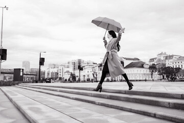 A girl in a light brown coat and a light scarf under an umbrella in a jump shot on a cloudy April...