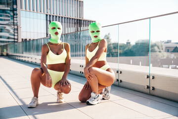 Two beautiful sexy women in green underwear. Models wearing bandit balaclava mask. Hot seductive female in nice lingerie posing in the street at sunny day, blue sky. Crime and violence. Perfect body - obrazy, fototapety, plakaty