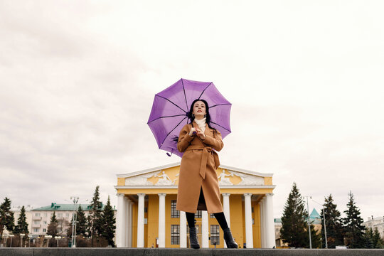 A girl in a light brown coat and a light shawl under a purple umbrella shot on a cloudy April evening, against the background of the Chuvash Drama Theater