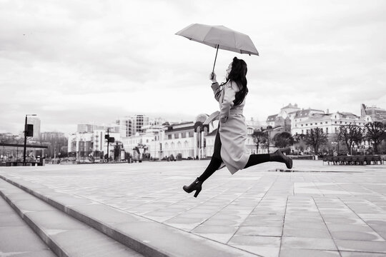 A girl in a light brown coat and a light scarf under an umbrella in a jump shot on a cloudy April evening against the background of Cheboksary