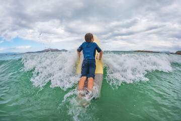 Happy boy take wave in surfing - young surfer learn to ride on surfboard with surf instructor on sea waves. Active family lifestyle, kids outdoor water sport lessons.