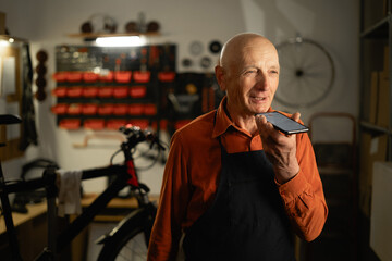 Old male bicycle mechanic doing his professional work in workshop using voice assistant on mobile...