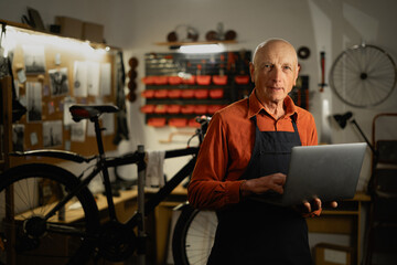 Portrait of a old repairman in workwear holding laptop computer at the bicycle workshop.