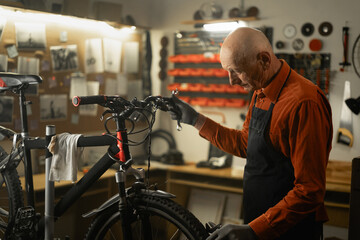 Bicycle repair in workshop. Old male mechanic working in bicycle repair shop, repairing bike in his garage - obrazy, fototapety, plakaty