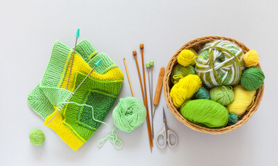 Top view of wicker basket with balls, skeins of green and yellow wool yarn, knitting needles and striped handmade knitted fabric. Hand knitting square pattern using 10-stitch method. Flat lay, closeup