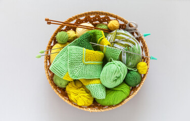 Top view of wicker basket with needlework: balls and skeins of green and yellow yarn, knitting needles and striped handmade knitted fabric. DIY concept. Flat lay, copy space, top view, closeup, mockup