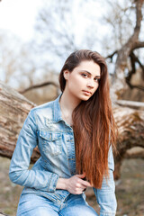 Naklejka premium young beautiful woman with brunette hair in a fashion jeans jacket sits in the park