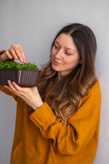 Woman in a mustard blouse admires a lush pot of microgreens, reflecting a moment of home gardening satisfaction. Arugula cultivation at home, fresh healthy greens rich in vitamins.