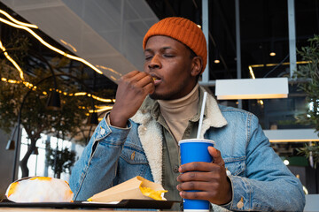 A young African American man enjoys fast food in a modern food court. A man in a trendy orange hat enjoys fast food, savoring French fries and holding a glass of soda in his hands