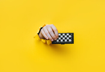 A child's hand emerges from a torn hole in yellow paper and holds a small chessboard. The concept...