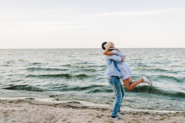 Happy and funny couple in love hugging on seashore. Female and male standing on beach ocean and enjoying sunny summer day. Man and woman embrace on sand sea. Cheerful couple spending time together.