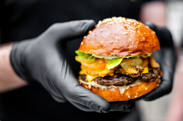 Close-up of a person in black gloves holding a juicy burger with a glossy bun, fresh lettuce, melted cheese, and a grilled patty against a blurred background