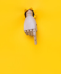 A wooden hand protrudes from a torn hole in yellow paper and points its index finger downwards. Concept of direction, adjustment and assistance. Mannequin, robot.