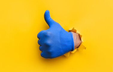 A left man's hand in a blue fabric work glove shows a thumb up (like). Torn hole in yellow paper....
