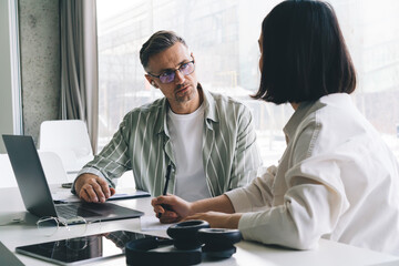 Fototapeta premium Male employee in optical spectacles for vision protection communicate with female colleague during cooperation process at table desktop with laptop computer, professional entrepreneurs talking