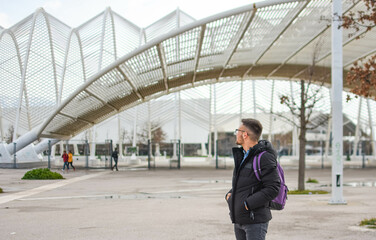 Young man in black jacket and purple backpack admires Athens Olympic complex architecture in background.