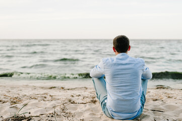 Young traveler man sits on white sand, enjoying amazing view. Back view. Handsome man walks near...