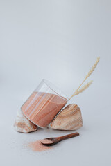 a glass jar with pink Himalayan salt stands at an angle on stones on a white background, next to a...