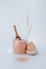a glass jar with pink Himalayan salt stands on a white background with two spikelets and a small wooden spoon stuck in the salt, next to it is a stone and the salt is scattered