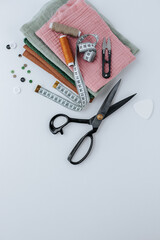a stack of different fabric scraps lies on a white background, on top are two skeins of thread, tailor scissors and a measuring tape. buttons nearby. shot from above
